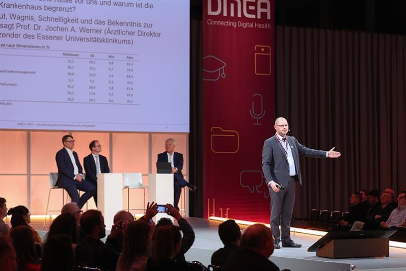Ein Mann spricht auf einer Bühne, hinter ihm sitzen drei weitere Speaker. Rundum ist Publikum zu sehen. Ein Mann spricht auf einer Bühne, hinter ihm sitzen drei weitere Speaker. Rundum ist Publikum zu sehen.