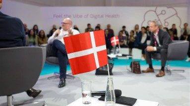 Im Hintergrund ist eine Talkrunde mit drei Männern zu sehen. Im Vordergrund steht eine kleine dänische Flagge auf einem Tisch.