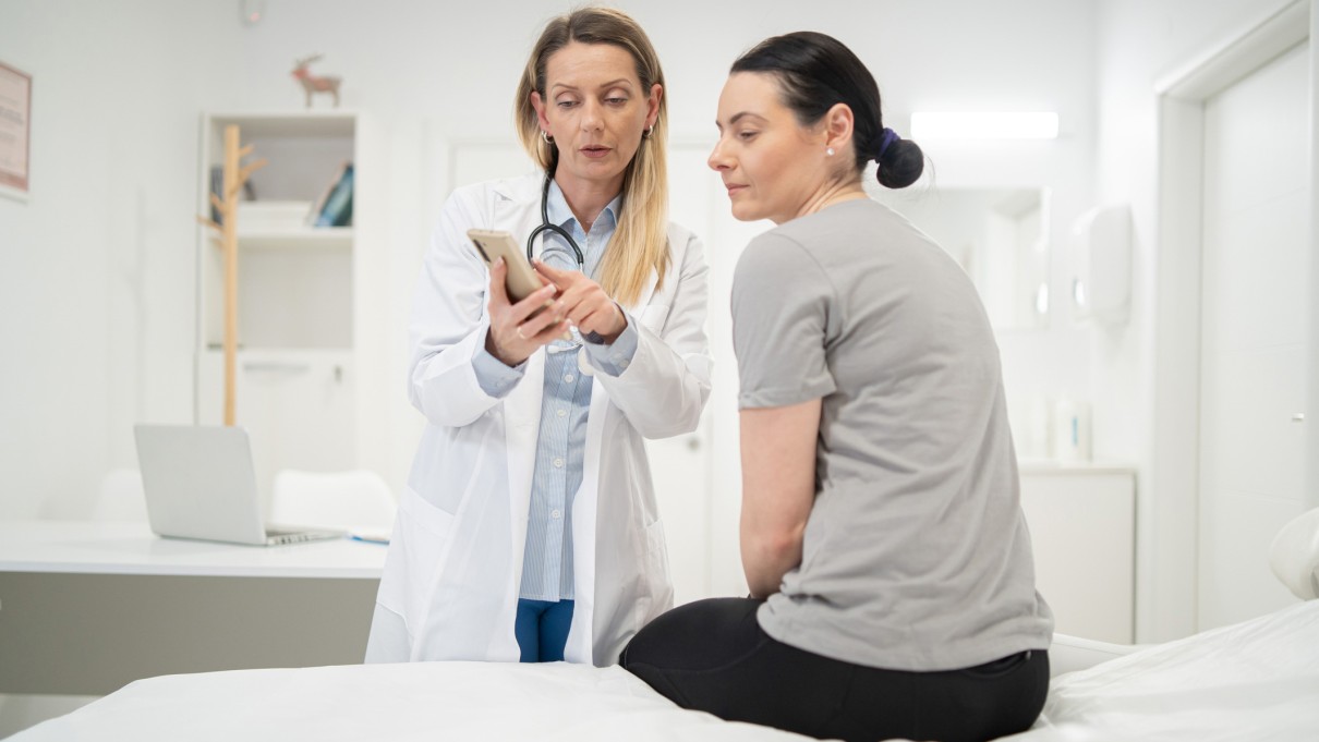 A doctor holds a tablet in her right hand and shows her patient something on the screen with her other hand.