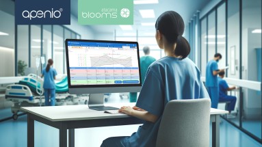 A nurse sits at a screen and looks at a graphic. Other hospital staff can be seen at work in the background.