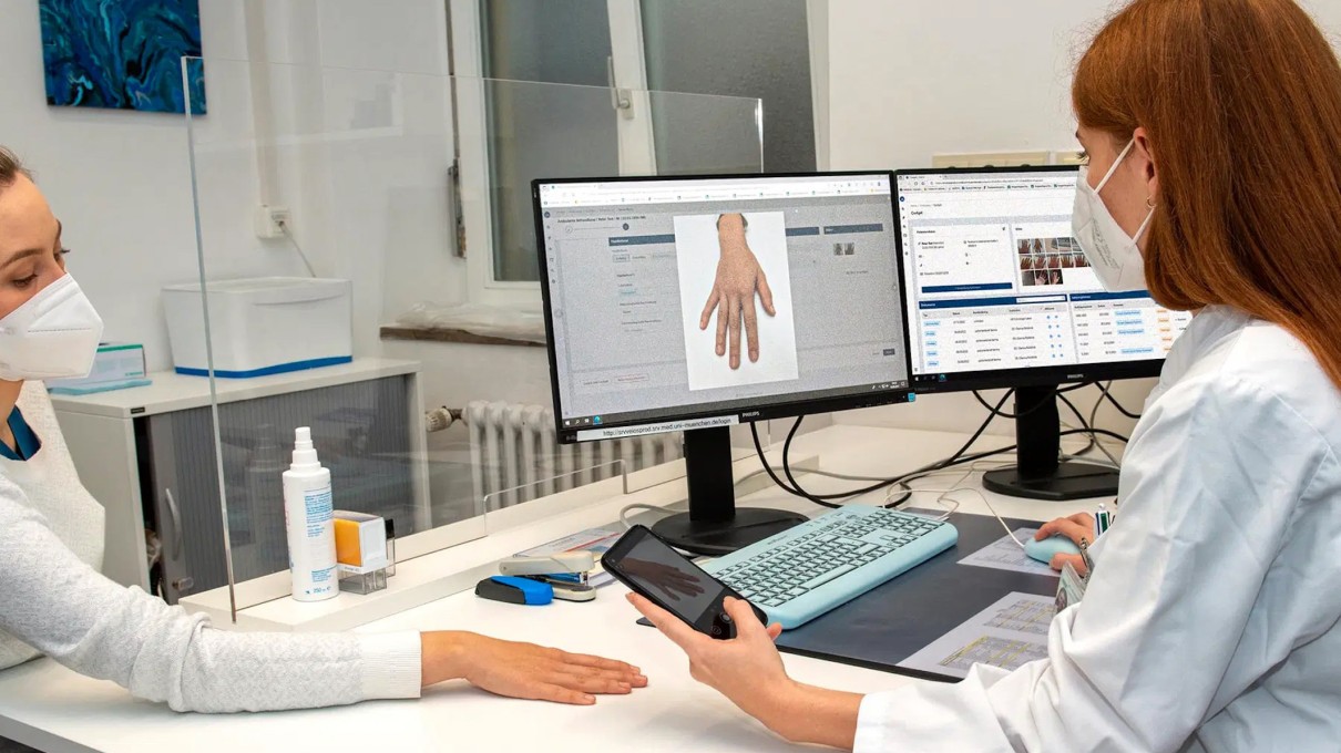 A doctor scans a patient's hand using a smartphone. The scanned hand can be seen on a screen.