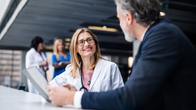 Das Bild zeigt eine Gesprächssituation zwischen einer Ärztin mit einem Patienten, im Hintergrund weiteres medizinisches Personal