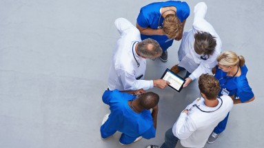 : Eine Gruppe mit Klinikpersonal steht um ein Tablet und berät über einen Befund, Blick von oben.