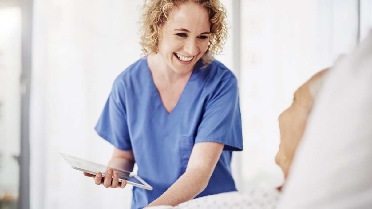 Eine Krankenschwester steht mit einem Tablet in der Hand am Bett einer älteren Patientin.