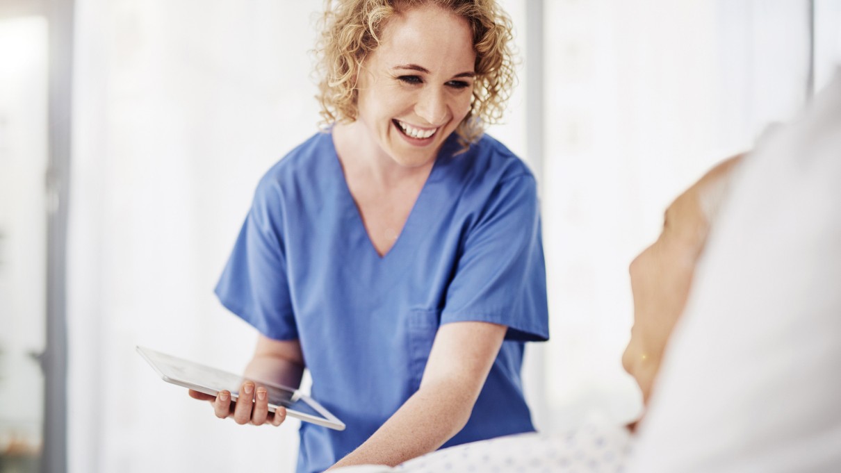 A nurse stands at the bedside of an elderly patient with a tablet in her hand