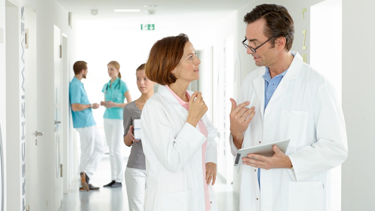 Female doctor talking to male doctor, practice staff in the background