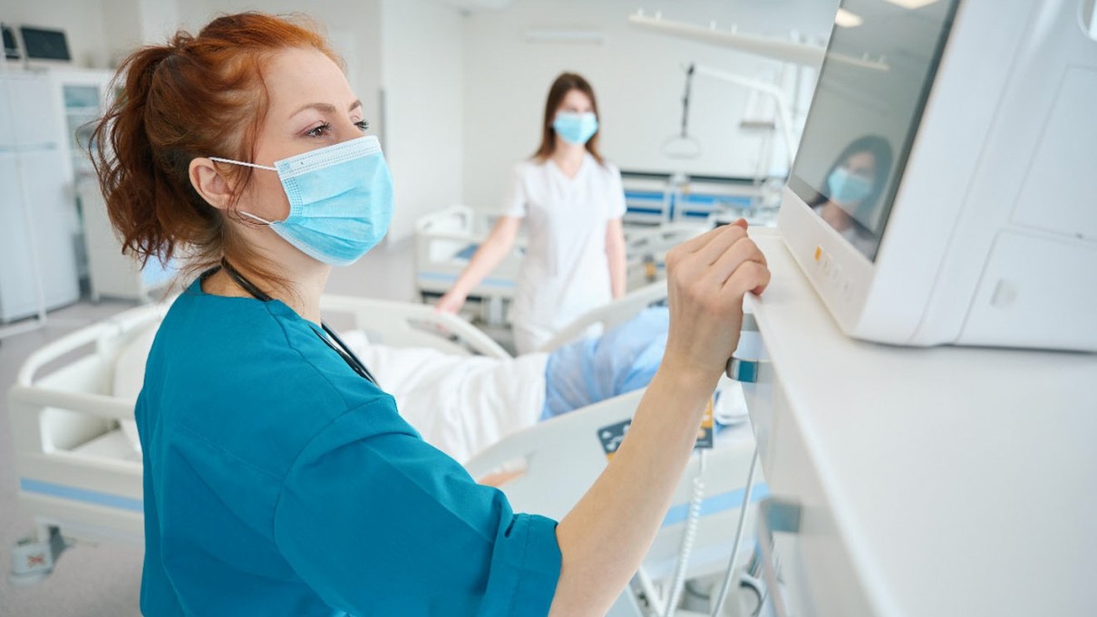 Krankenschwester sieht auf einen Bildschirm, Patientenbett und andere Krankenschwester im Hintergrund
