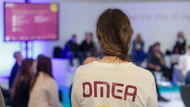 Eine Frau mit geflochtenen Haaren von hinten, auf dem weißen Pulli ist das DMEA-Logo zu sehen.