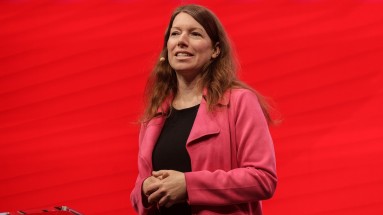 A woman with long brown hair is speaking on stage. She is wearing a pink jacket over a black top and a headset microphone. 