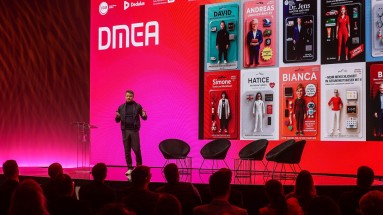 A speaker is standing on a stage at the DMEA in front of a large red background. In the foreground, the audience can be seen.