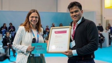 Presenter on the left with microphone and DMEA sparks card next to Minh Duc Do, who is holding the certificate for second place 