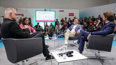 Panel discussion on a stage with four people seated in armchairs, engaged in conversation. 