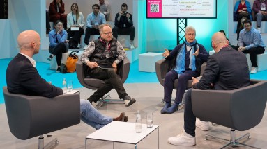 Discussion with four participants on a stage at DMEA 2025, surrounded by an audience in a trade fair atmosphere. 