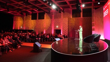 A woman in a green pantsuit stands on a circular stage, speaking to an audience at a conference. 