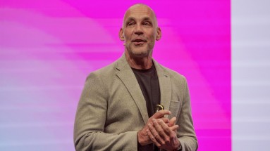 A bald man with a grey beard stands on a stage with a magenta background. He is wearing a beige jacket.