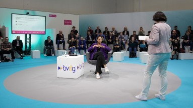 A woman stands with notes in her hand in front of a seated panelist, surrounded by an audience on white seating cubes.