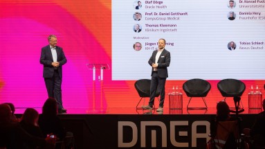Two men in suits stand on a stage at the DMEA in front of a large screen.