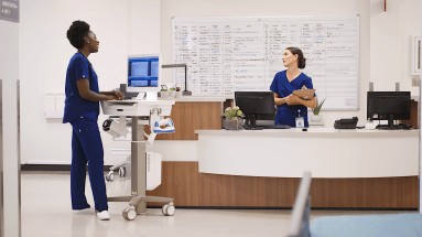 Two nurses talking. The one on the left is standing at an Ergotron CareFit cart, the one on the right is standing at a computer.