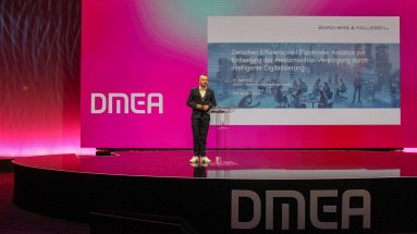 A man in a suit stands on a stage at the DMEA in front of a large screen.